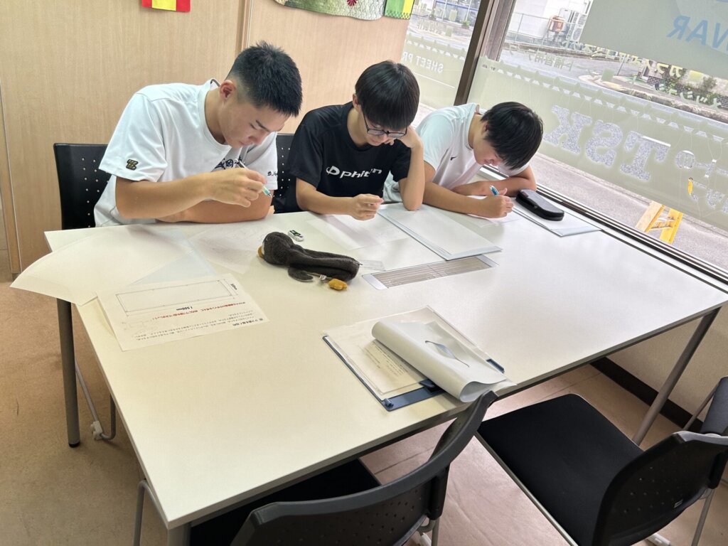 Three young men are intently focused on their studies around a white table, surrounded by papers and books. They appear to be engaged in a learning or work session.