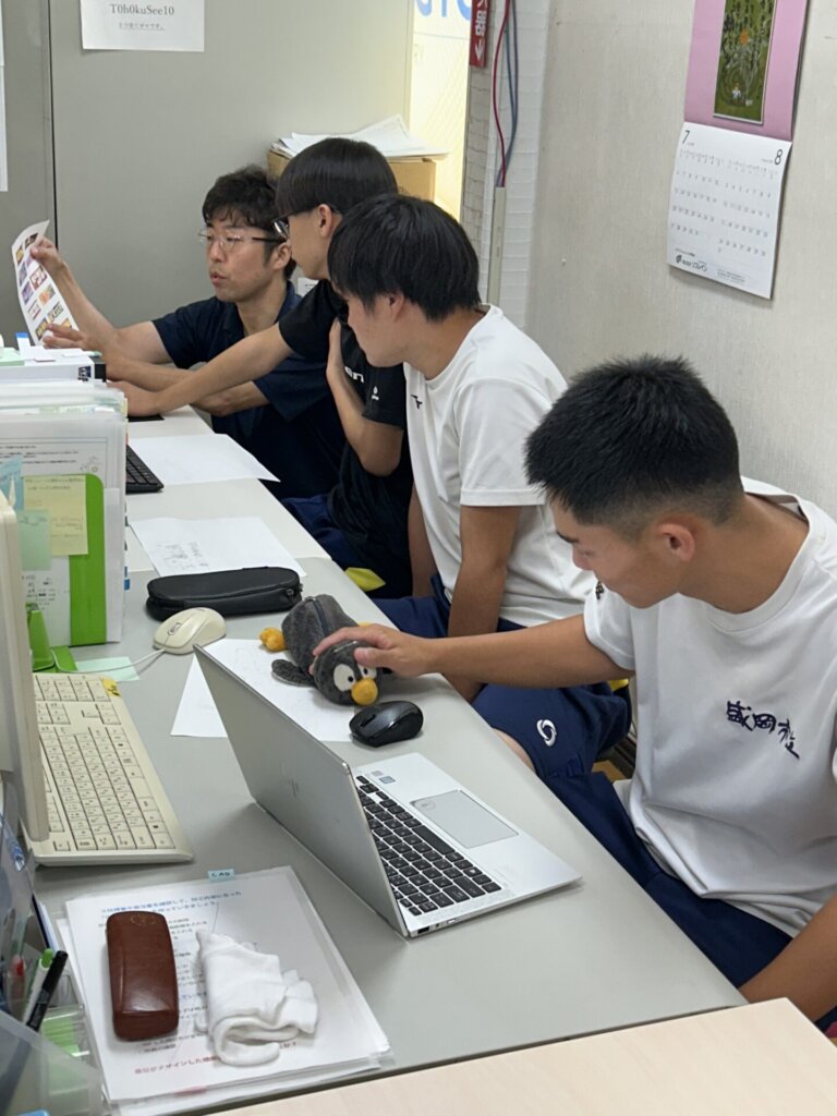 Four young men are gathered around a desk, looking at documents and laptops. One is holding up a paper while another interacts with a penguin-shaped object on a laptop, possibly engaged in a study or work session.